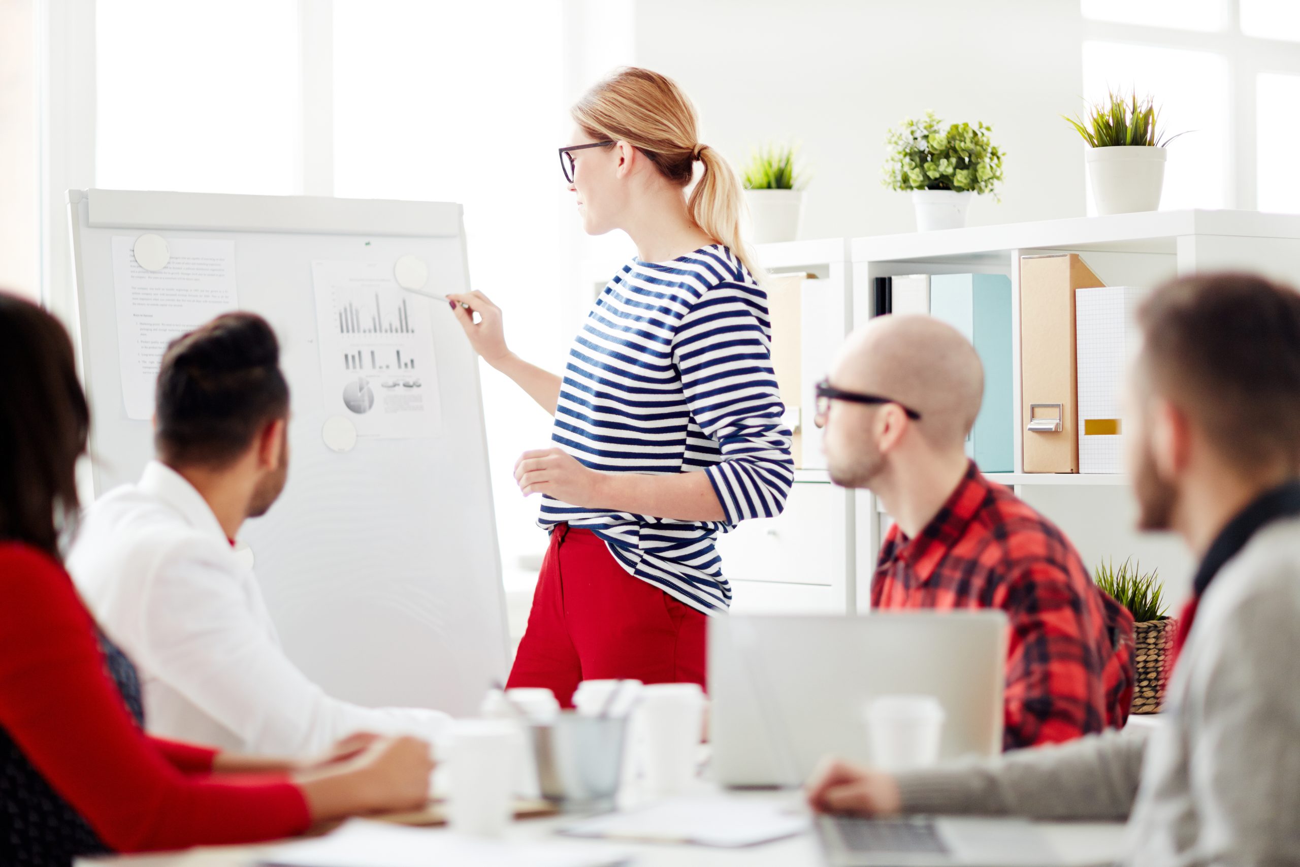 Business lady or teacher standing by whiteboard and pointing at financial document while group of people listening to her presentation